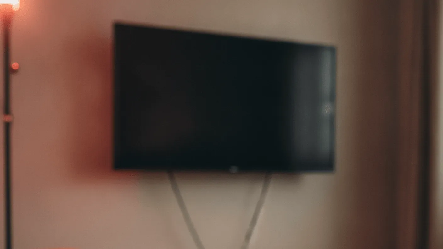 A man's hand rests on a remote control on a wooden table, with a wall-mounted television in the background.