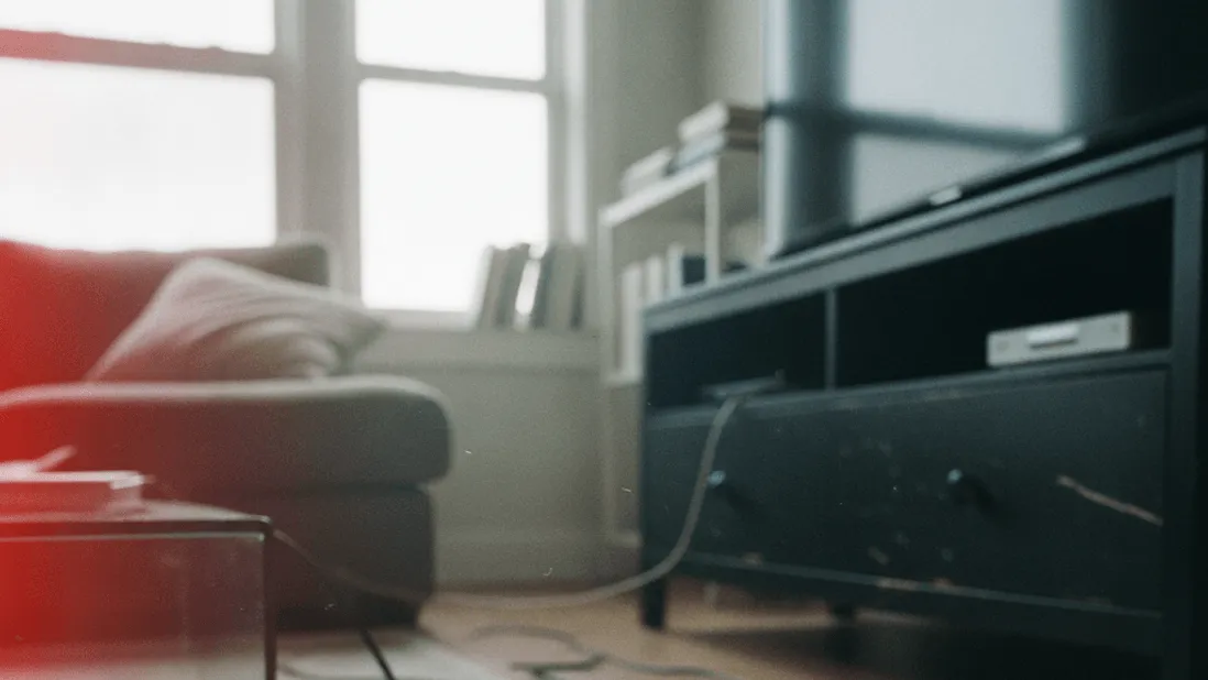 A hand holds a smartphone showing a clock app beside a television remote on a rug, midday light from a window.