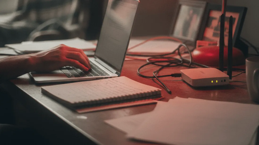 A hand rests on a laptop keyboard next to a notebook, under the red glow of a desk lamp.