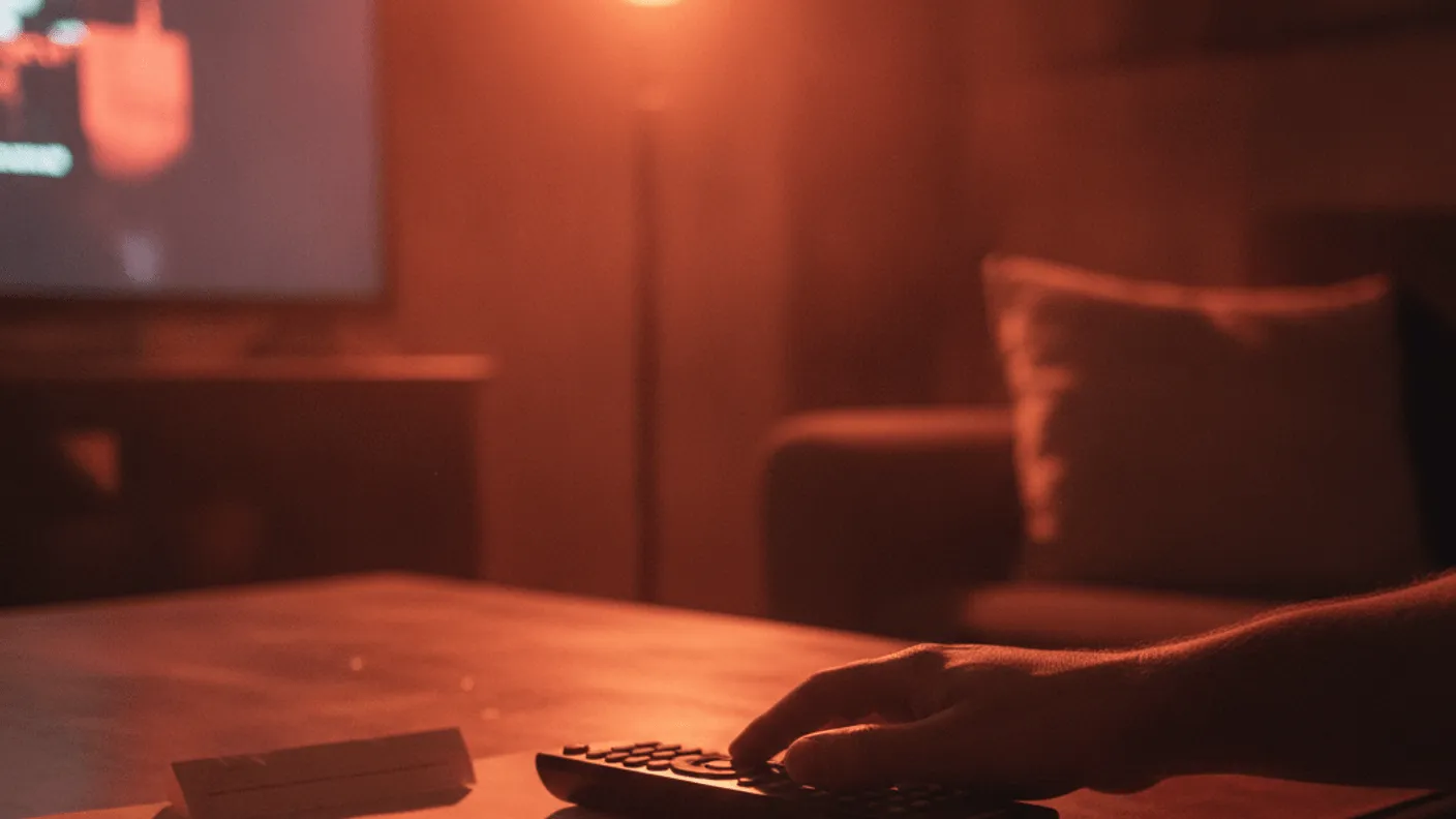 A hand rests on a television remote next to a checklist, illuminated by a lamp and a softly glowing television screen.