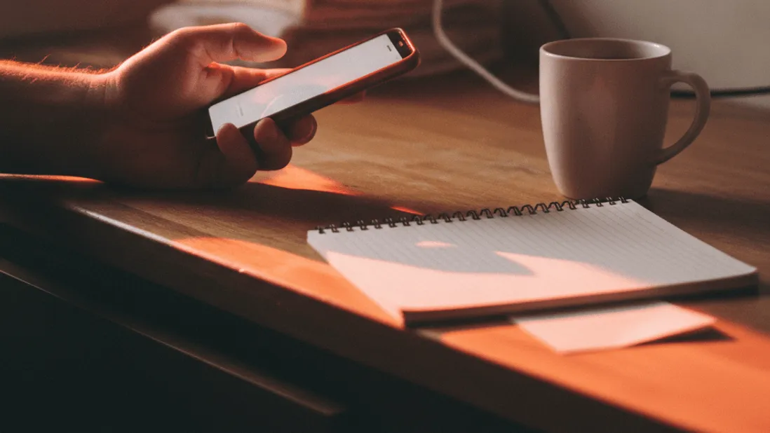 A hand holds a phone over a counter with a coffee mug and notebook nearby in soft morning light.